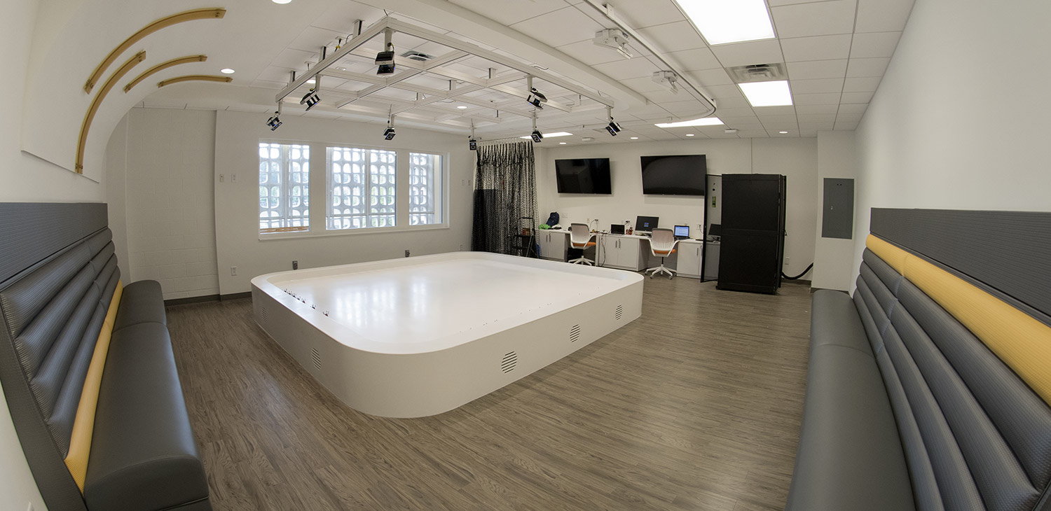 The robotarium, a sleek white room with a shallow vinyl pool in the middle which robots swarm in. Booths for viewers line the walls and a large window is on the far wall for passing audiences