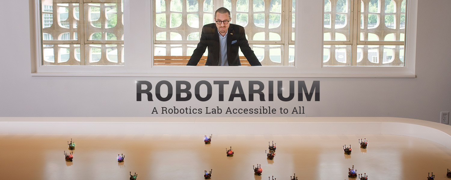 Magnus Egerstedt looking through a large viewing window into the Robotarium, which has a shallow white vinyl pool that small robots are swarming in