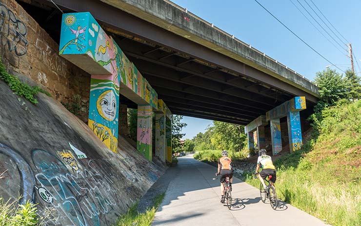 Two bicyclists pass beneath a bridge painted with murals