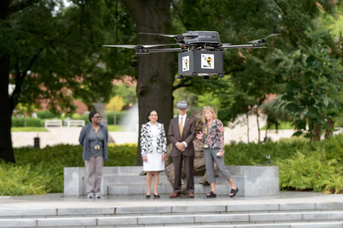 A drone with a box underneath flying about ten feet off the ground with people observing