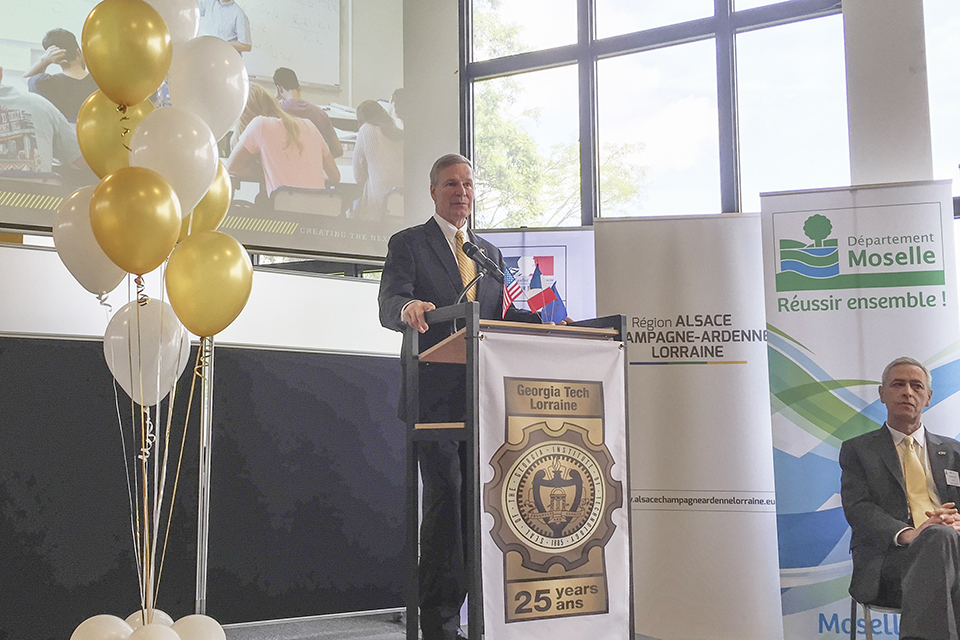 President Peterson speaking at a podium with a GT slideshow and balloons in the background. Yves Berthelot is sitting at the side of the stage.