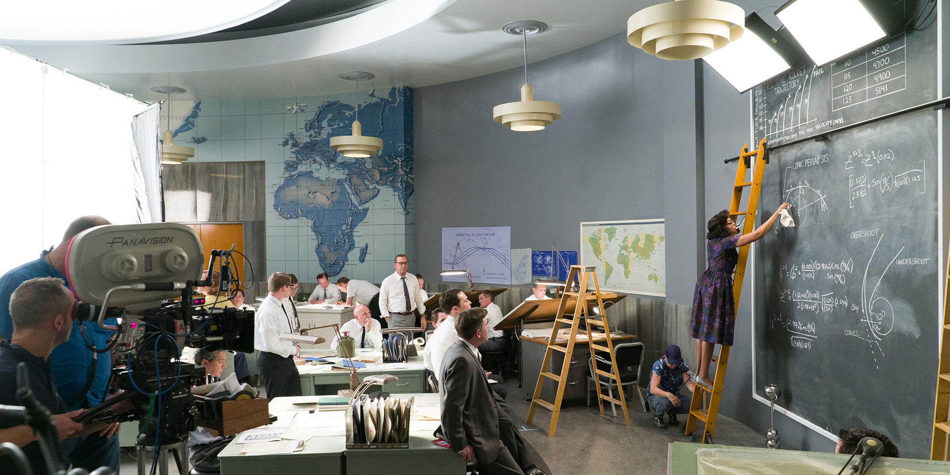 1960s style office with large blackboard on one wall, woman writing equations on board as men sit and watch