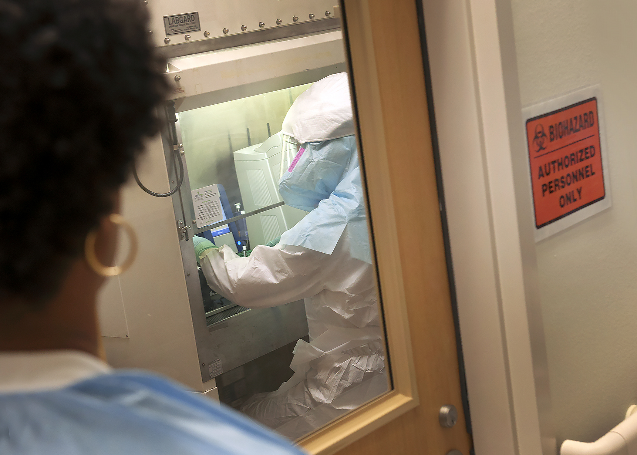 Image shows a lab technician in Emory’s Serious Communicable Diseases Unit laboratory, with a team member watching from the outside for safety purposes. (Credit: Jack Kearse, Emory Health Sciences Photography)